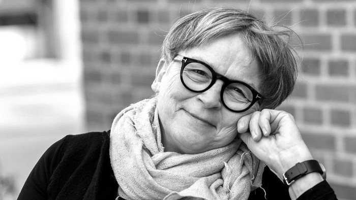 En glad kvinde med briller og et tørklæde, der støtter hovedet på hånden udendørs.