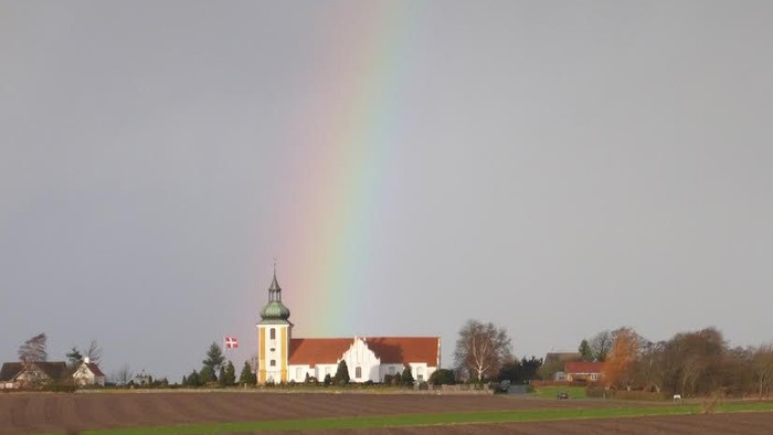 En regnbue over en åben mark med en kirke i baggrunden