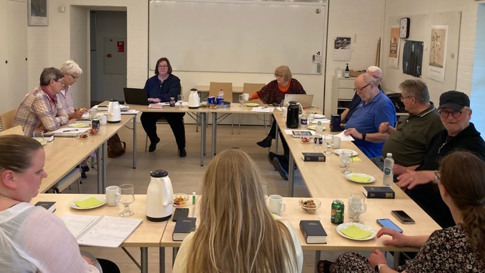 En gruppe af mennesker sidder ved lange bord og spiser morgenmad i et lys og rummeligt lokal.