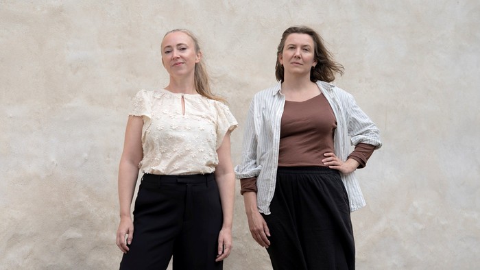 To kvinder poser selvsikkert foran en tekstureret væg i stilfuldt, afslappet tøj.