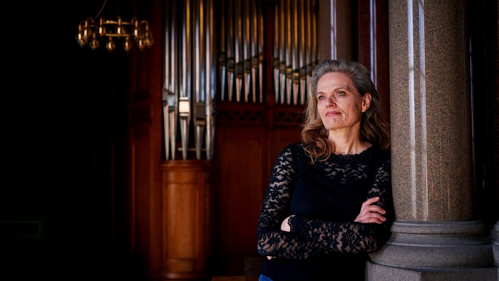 En kvinde står selvsikkert ved siden af et prægtigt pibeorgel i en svagt oplyst kirkes indre.