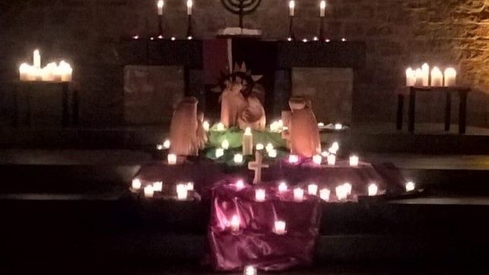 Im dunklen Raum steht ein Tisch mit vielen Kerzen und einem großen Blumenstrauß, darüber hängt ein großes Kreuz.