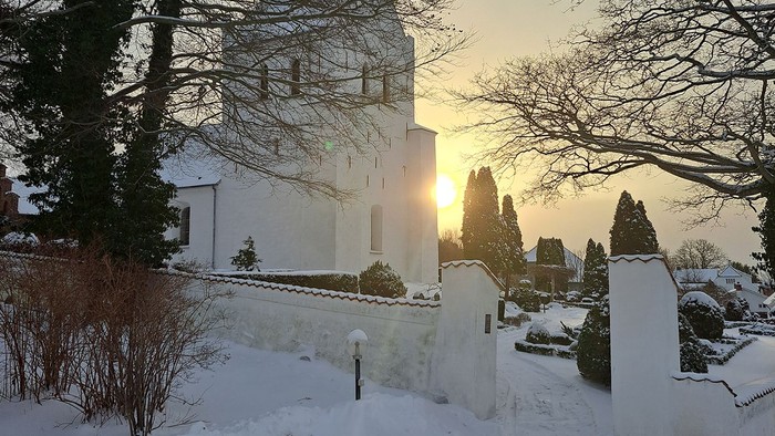 En fredfyldt vinterscene med en hvid bygning badet i gyldent solnedgangslys gennem sneklædte træer.
