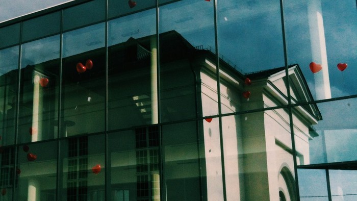 Modernes Glasgebäude mit roten Herzdekorationen, das umliegende Strukturen und den Himmel widerspiegelt.