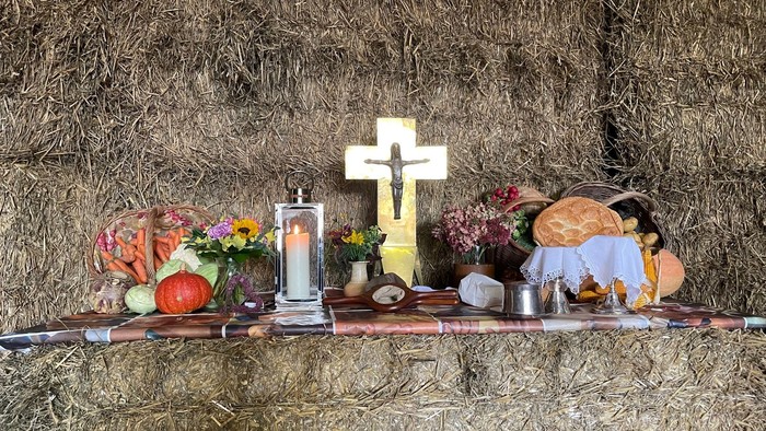 Ein rustikaler Altar mit Essen, Blumen, einer Kerze und einem Kreuz auf einem mit Heu bedeckten Hintergrund.