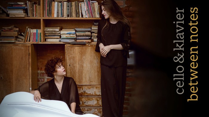 Eine Frau im Bett blickt zu einer anderen Frau auf, die neben ihr steht, mit Bücherregalen im Hintergrund.