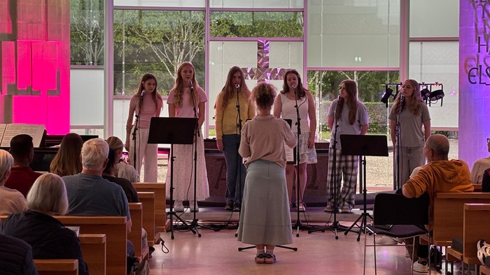 En gruppe sangere optræder på scenen i et stærkt oplyst, moderne kapel med et opmærksomt publikum.