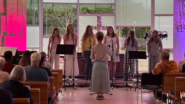 En gruppe sangere optræder på scenen i et stærkt oplyst, moderne kapel med et opmærksomt publikum.