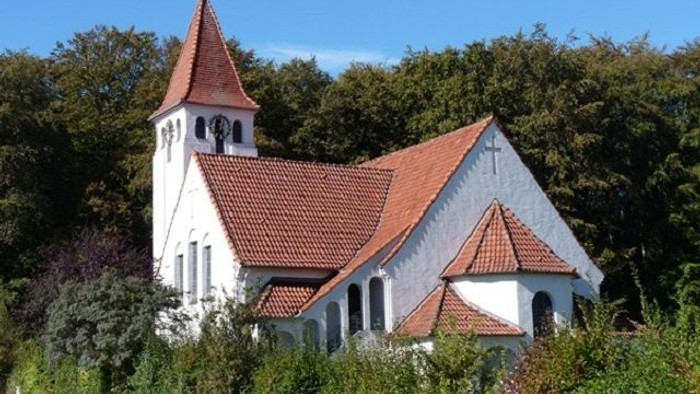 Weiße Kirche mit rotem Dach und Turm, umgeben von Bäumen und Büschen