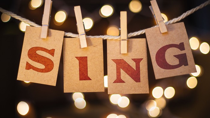 Braune Papierkarten bilden mit Wäscheklammern das Wort „SING“ und hängen vor einem unscharfen Lichterhintergrund.