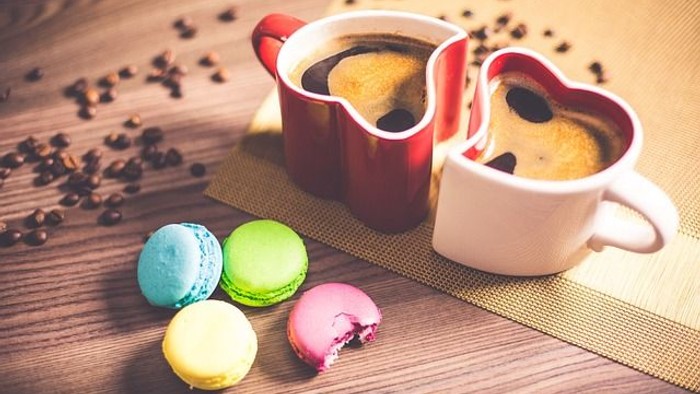 Zwei herzförmige Tassen mit Kuchen und bunte Macarons auf einem Holztisch