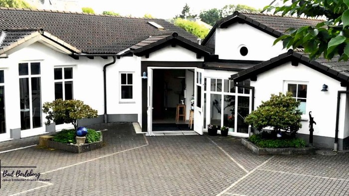 Ein charmantes weißes Gebäude mit einem zentralen Eingang, umgeben von gestalteter Grünanlage und einem gepflasterten Innenhof.