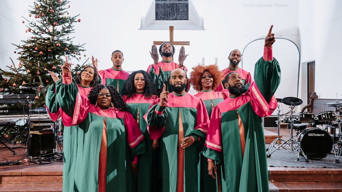 Ein Kirchenchor in leuchtend grün-roten Gewändern singt vor einem Weihnachtsbaum und einem Kreuz.