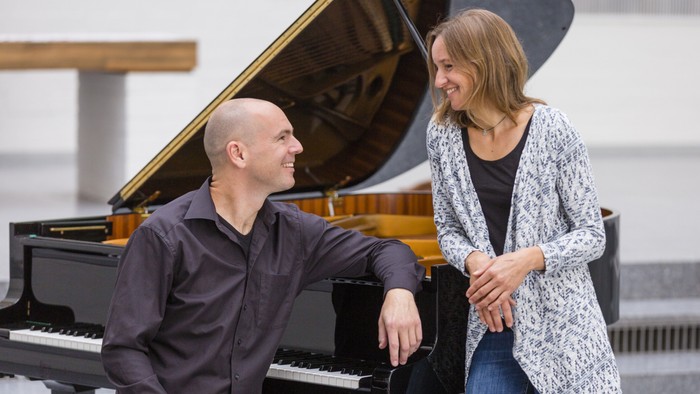 Ein Mann sitzt an einem Flügel und lächelt eine Frau an, die neben ihm steht.