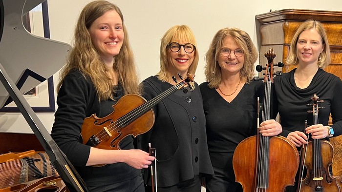 Vier Musiker posieren gemeinsam mit Streichinstrumenten und einem Flügel in einem gemütlichen Raum.