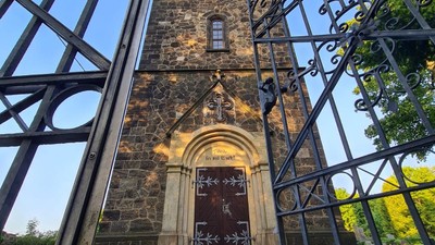 Eisenzaun vor alter Steinmauer mit Türmchen und Kreuz.