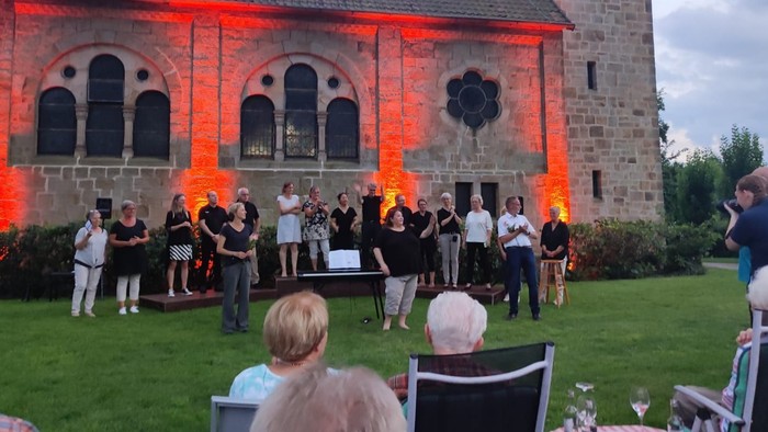 Vor der angestrahlten Johanneskirche sieht man einen Chor auf der Wiese stehen, im Vordergrund sitzen Zuhörer an Picknicktischen.