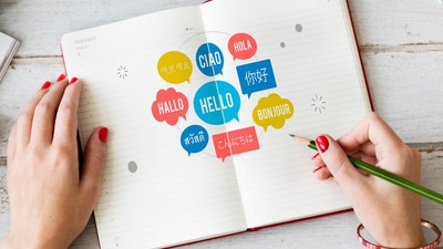 Eine Person schreibt in ein aufgeschlagenes Notizbuch mit bunten Sprechblasen und Begrüßungen in verschiedenen Sprachen.