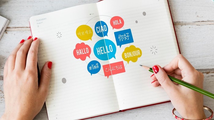 Eine Person schreibt in ein aufgeschlagenes Notizbuch mit bunten Sprechblasen und Begrüßungen in verschiedenen Sprachen.