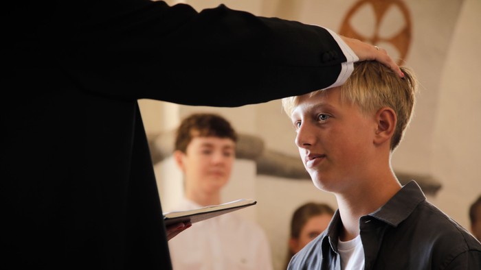 En ung dreng med blond hår står med en hånd på sit hoved og ser til siden.