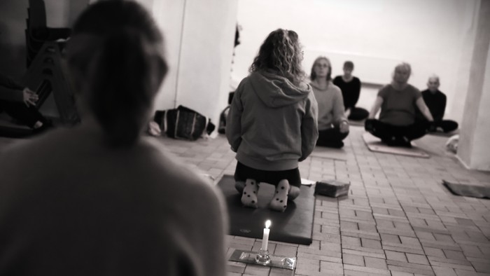 En gruppe af mennesker sidder på yoga-matter i en rum.