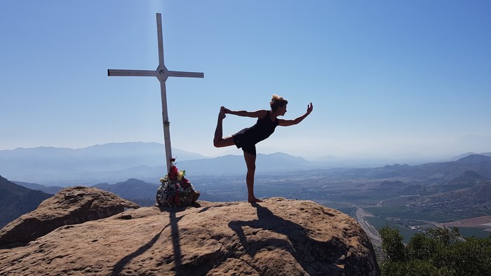 En person udfører yoga på en klippe ved et kors med udsigt over bjerge og dal.