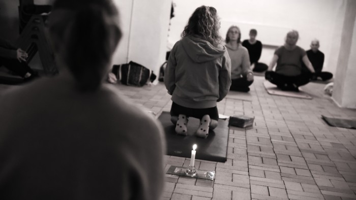 En gruppe af mennesker sidder på yoga-matter i en rum.