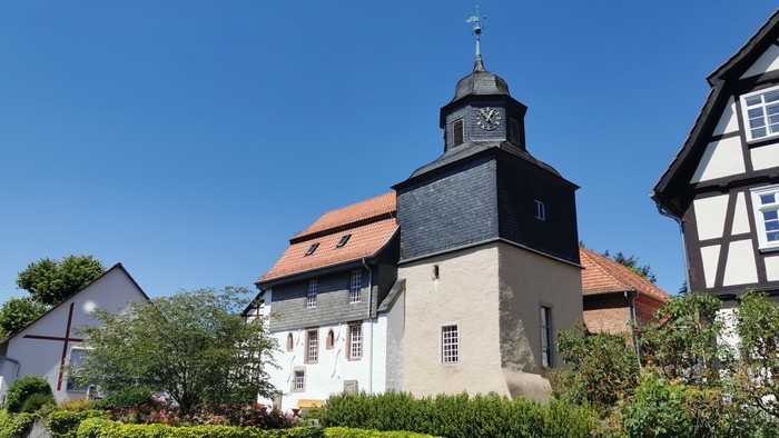 Das Bild zeigt eine Kirche mit schwarzem Dach und weißem Turm, umgeben von grünen Büschen und anderen Häusern.