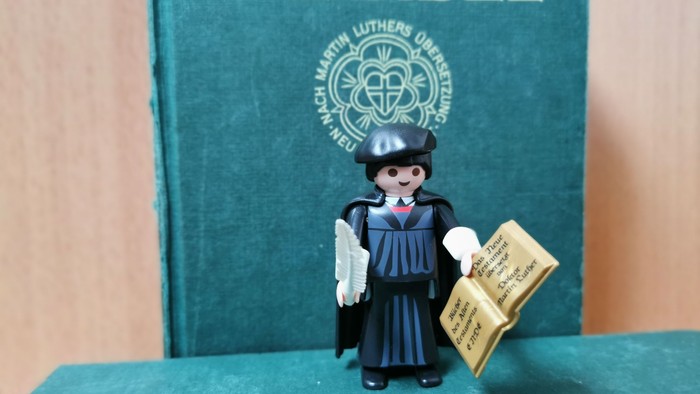 Eine Playmobil-Figur in Mönchsgewand hält ein Buch vor einem grünen Buch mit der Aufschrift