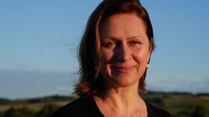 Eine Frau mit kurzen braunen Haaren lächelt im Freien vor einer malerischen, weiten Landschaftskulisse.