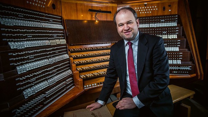 Ein Mann in formeller Kleidung steht neben einer großen, kunstvollen Pfeifenorgel-Konsole.