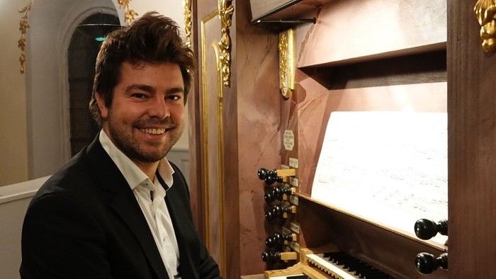 Ein Mann in formeller Kleidung sitzt lächelnd neben einer verzierten Pfeifenorgel.