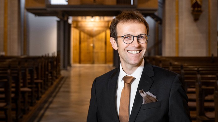 Ein Mann im Anzug steht lächelnd vor einer verzierten Kirchenorgel und Kirchenbänken.