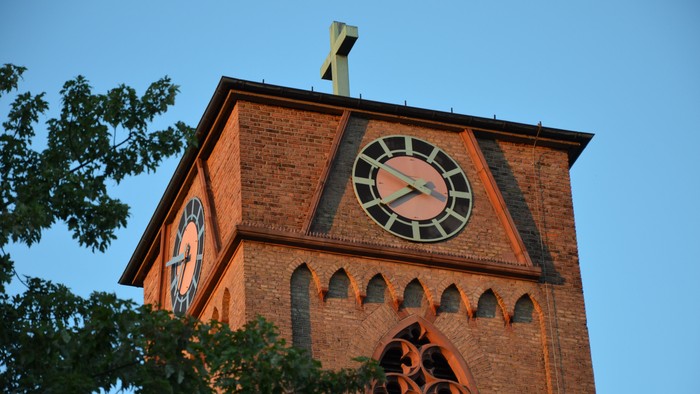 Der Kirchturm aus Ziegeln mit Uhr und Kreuz.