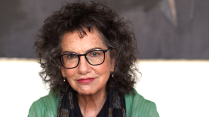 Eine selbstbewusste Frau mit lockigem Haar trägt eine Brille, eine grüne Jacke und einen gemusterten Schal und sitzt nachdenklich da.