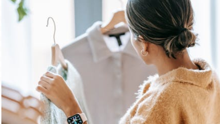 Eine Frau in einem gemütlichen Cardigan betrachtet Kleidung, die an einer Kleiderstange im Schrank hängt, während sie eine Smartwatch am Handgelenk trägt.