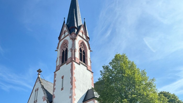 Weiße Kirche mit hohem Turm und spitzen Dach, umgeben von Bäumen und einem klaren blauen Himmel.