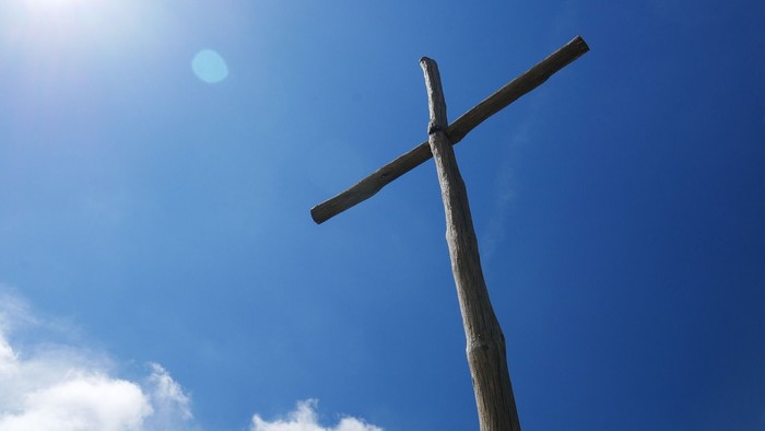Ein hölzernes Kreuz vor einem klaren blauen Himmel mit einigen Wolken und der Sonne.