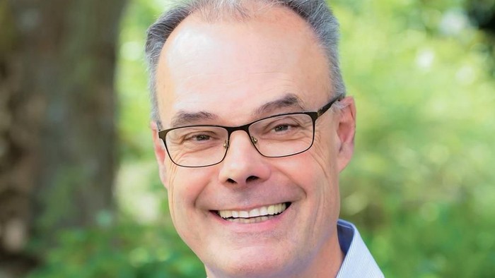Ein lächelnder Mann mit Brille steht im Freien vor einem grünen, blättrigen Hintergrund.