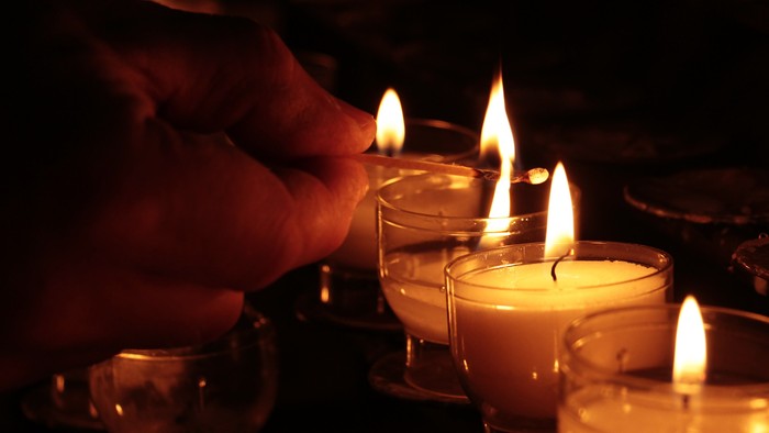 Eine Hand, die mehrere Kerzen in schwach beleuchteten Glasbehältern anzündet und warmes, flackerndes Licht wirft.
