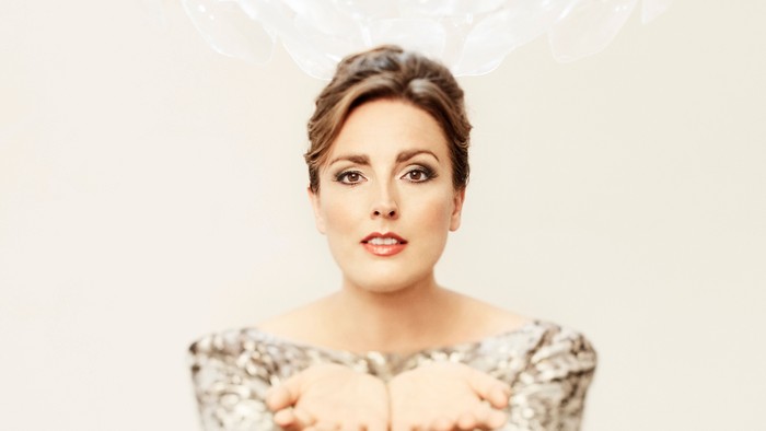 En kvinde med hår i en opstyling, der holder hænderne op forans, og har et spående udtryk.