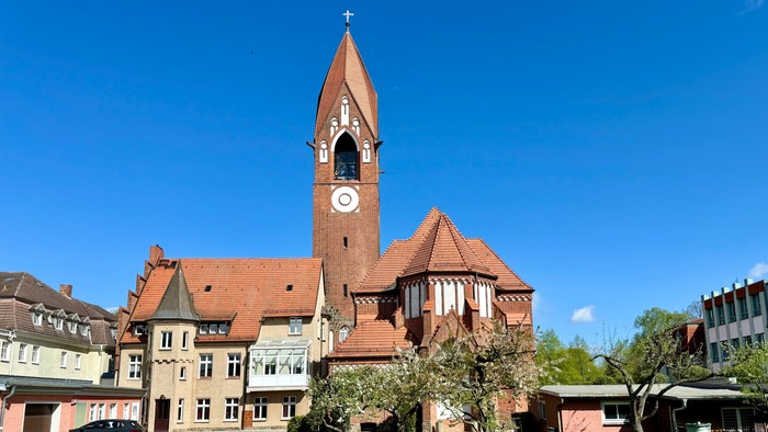 Bild zeigt eine Kirche mit hohem Turm und Uhr, umgeben von einem gepflegten Garten.