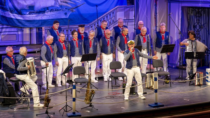 Gruppe von Männern in Matrosenoutfits, die mit Mikrofonen und Notenständern auf der Bühne auftreten.