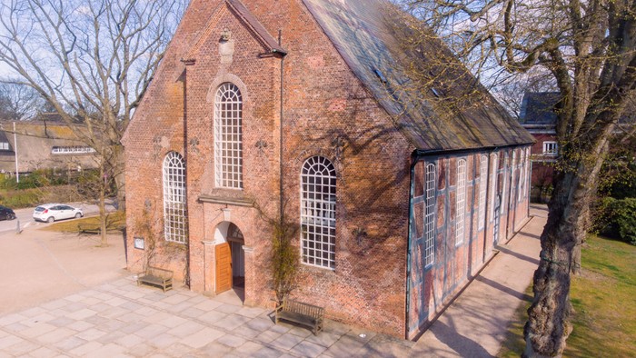 Kleines Backsteingebäude mit spitzen Fenstern und Tür, umgeben von Bäumen und einem gepflasterten Weg