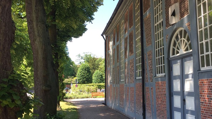 Links ein großer Baum, rechts ein langes, rotes Backsteingebäude mit weißen Fenstern und Türen.