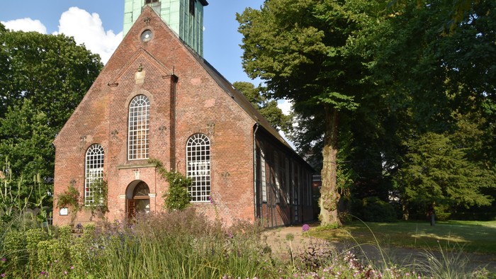 Ein altes Backsteingebäude mit Uhrturm und großen Fenstern, umgeben von üppigem Grün und einem Teich.