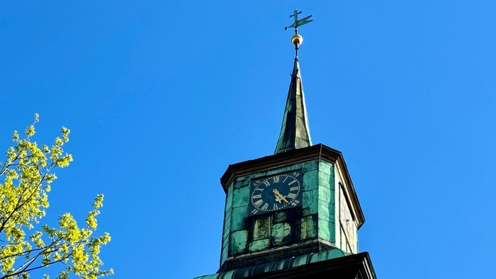 Ein historischer Uhrturm mit grüner Patina und einer spitzen Turmspitze vor einem klaren blauen Himmel.