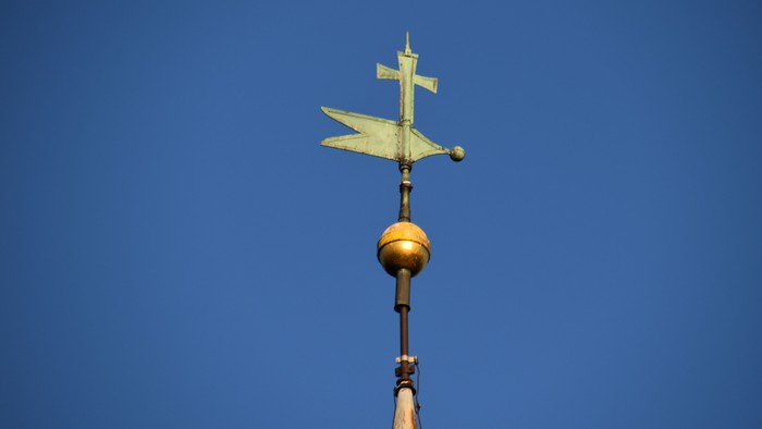 Ein goldener Wetterhahn in Form eines Flugzeugs auf einem verzierten Mast vor klarem blauem Himmel.