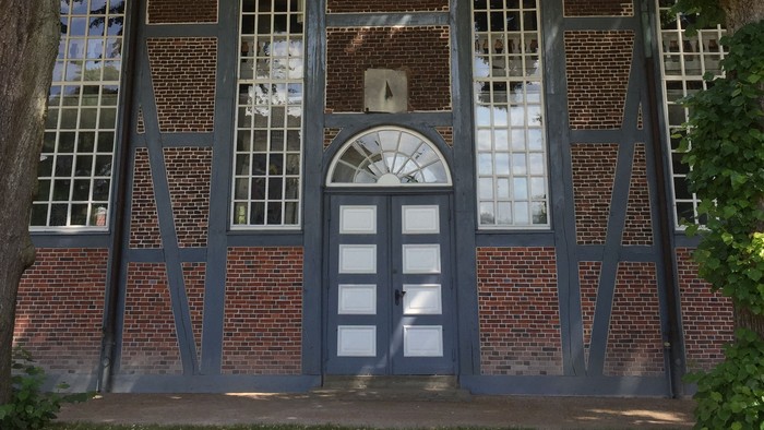 Historisches Backsteingebäude mit Fachwerk und Bogenfenstern, umgeben von Grün.