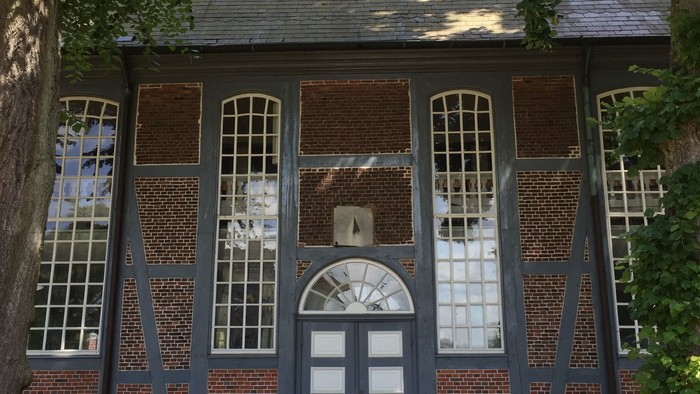 Historisches Backsteingebäude mit Fachwerk und Bogenfenstern, umgeben von Grün.